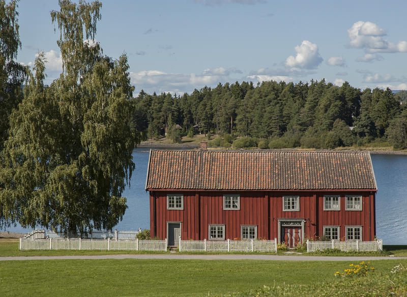 Stor, rød bygning i to etasjer ligger bak et hvitt stakittgjerde ned mot vannet. På venstre side to bjørker.