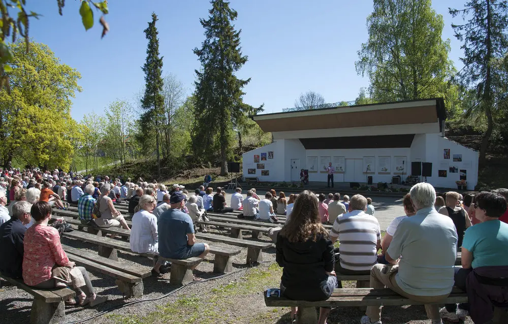 Friluftsscene med artister på og trebenker fylt med publikum