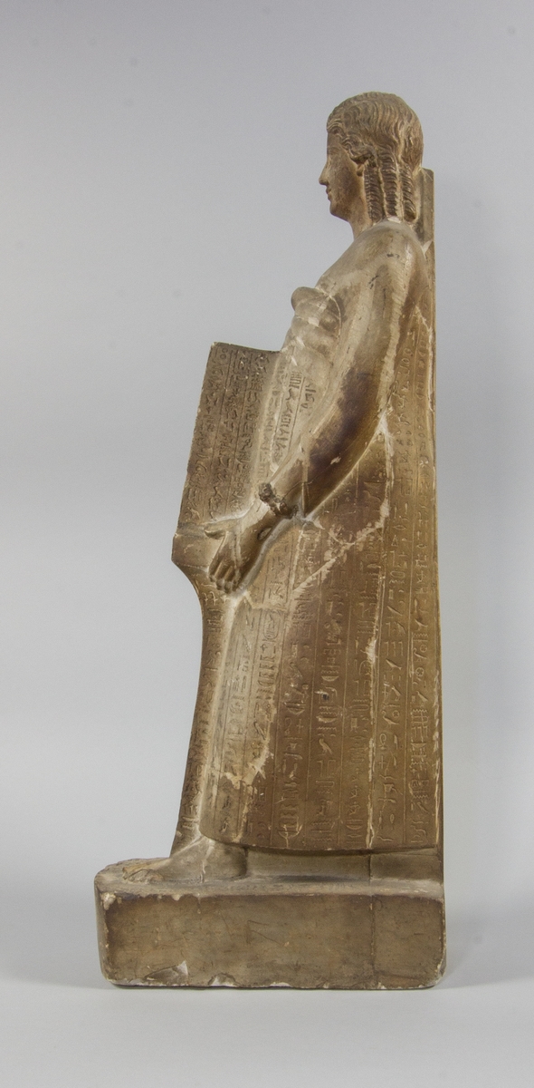 Kvinnogestalt stående i helfigur i en egyptisk pose med fötterna i en rak linje. Klädd i lång klädnad med bara axlar. Hon håller med båda händerna en stående rektagulärt öppen sarkofag innehållande en möjlig mumie av en farao. Ytorna täckta med hieroglyfer.