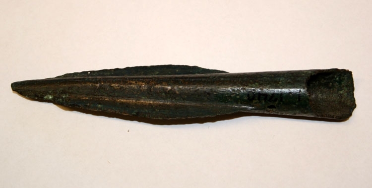 Spjutspets av brons med en 4 cm lång holk med nithål på båda sidor nedan det lancettformiga bladet. Holkens nedkant är skadad. Inskriven i huvudboken 1892-1900.