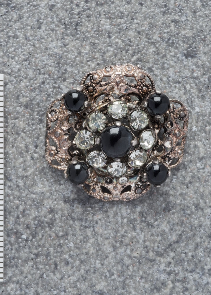 Smykkenål i sølv (antatt) med smykkesteiner. Firkløverformet med 5 sorte "perler" plassert som på en terning. Rundt den midterste perlen 8 diamantlignende steiner. På baksiden nål med festeanordning.