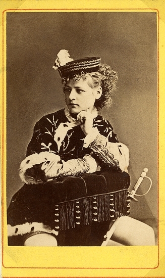 Rollporträtt (knäbild, halvprofil) av en kvinna i pagedräkt som sitter grensle över en stoppad stol.