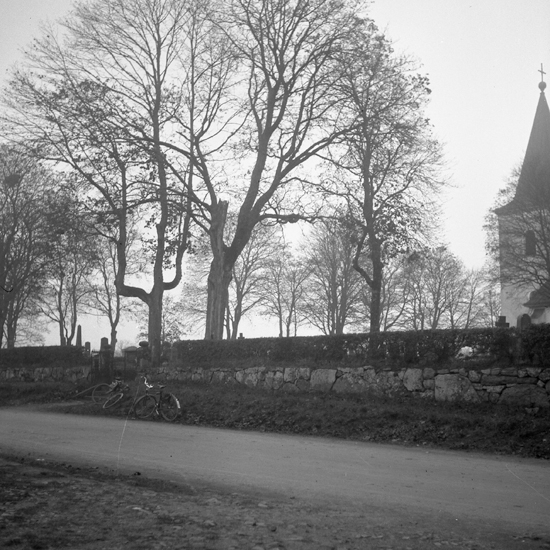 Berga kyrka, Lagan. I förgrunden syns en mindre grusväg vid stenmur och bakom syns kyrkogården.Till höger skymtar kyrkan.
