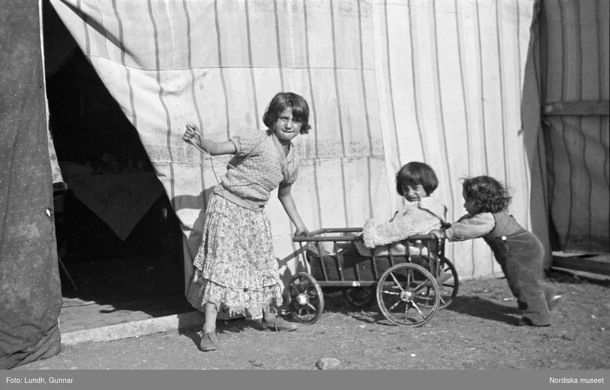Tre barn leker med en vagn/skrinda. Den äldsta flickan ser in i kameran. Familjen Taikons läger i Johanneshov, södra Stockholm.