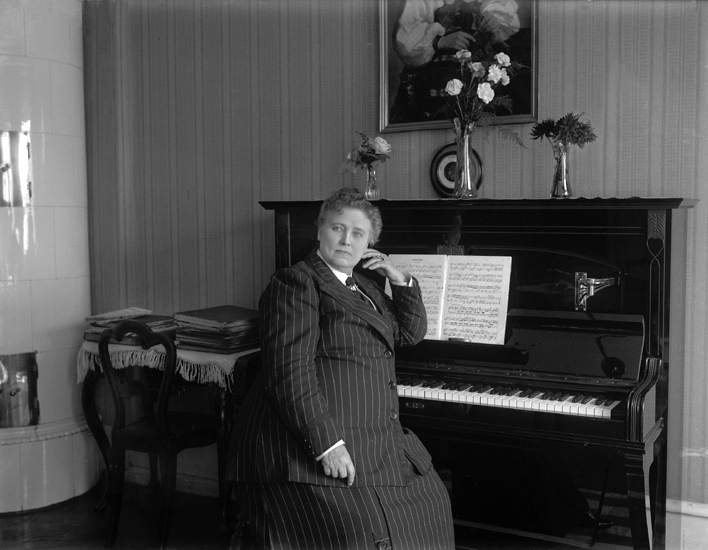 Christina Nilsson på sin födelsedag vid ett piano. Jämte pianot står ett bord med en stol.