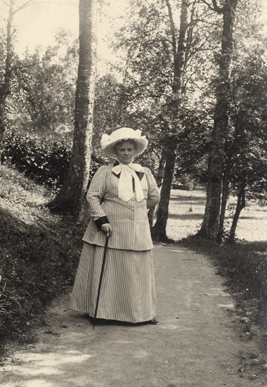 Porträttfoto av Christina Nilsson iförd promenaddräkt med hatt. Hon står på en mindre grusväg med träd runt omkring.