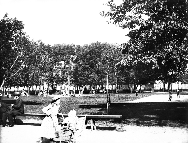 Foto från Societetsparken i Varberg. I förgrunden syns en flicka som drar en barnvagn och två herrar som sitter och diskuterar på en bänk. 
I bakgrunden till höger syns två pojkar som spelar badminton (?). 
Hårt, överexponerat foto.