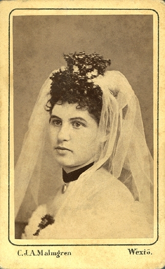Porträtt (bröstbild, halvprofil) av en brud i vit brudklänning, slöja och myrtenkrona m.m.. Runt halsen har hon ett mörkt sammetsband med en medaljong.