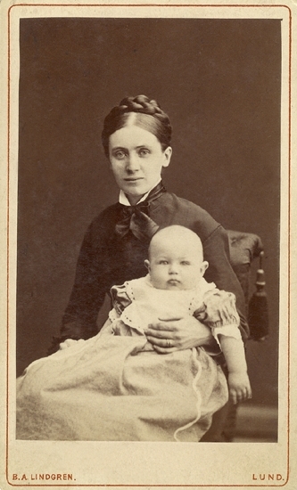 Porträtt (knäbild, halvprofil) av en okänd kvinna i mörk flätfrisyr och dito klänning, med en baby i famnen. Hon sitter på en stoppad stol, prydd med en tofs.