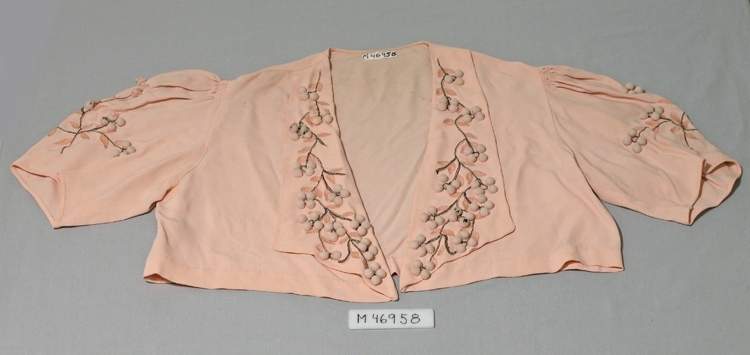 Långklänning av crepat rosa tyg. Ärmlös, smockrynk på axlar och ärmar.
Knäppes fram med 14 klädda knappar. Klockad nederdel, knytskärp, 1630 mm långt. 
Kort jacka med maskinsydd brodyr i rosa och brunt på ärmar och slag, 
samt klädda kulknappar.
Jackans längd 340 mm.


Inskrivet i huvudbok 1985