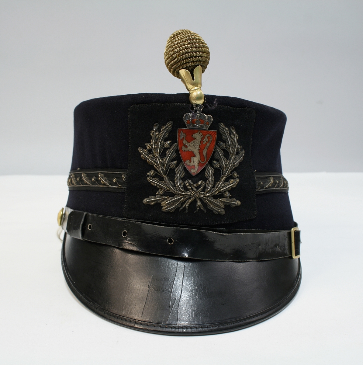 Mørkeblå kepi med blank skjerm med riksvåpen, ekeløvsdekor og pompon i gull.