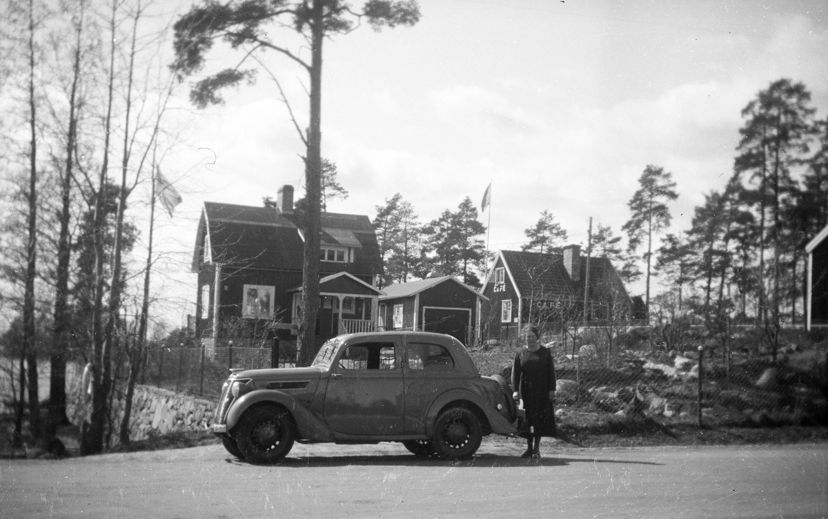 Bostadshus, kvinna vid en bil, en Ford Eifel 1939, och ett Café.