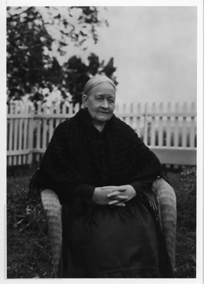 Eldre dame  med svart forkle og sjal sittende i en kurvstol. Damen er Pernille Lund f. Pernille Røsbakk.