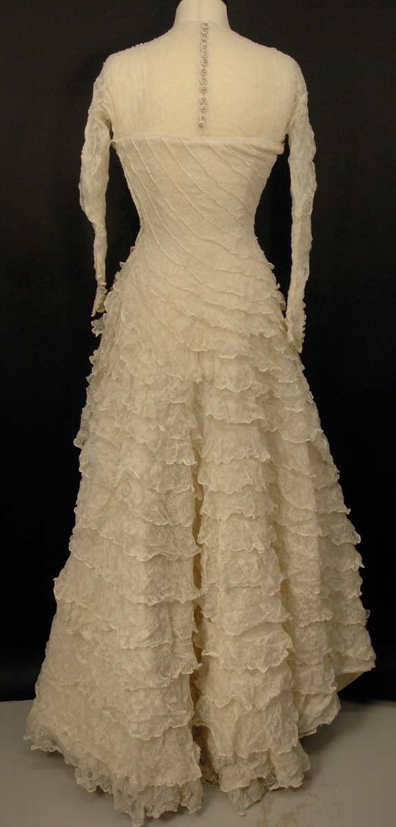 Helklipt, figursydd. Ermer og bærestykket klipt i ett. Rund hals bak med 22,5 cm lang splitt og 14 knapper med sydde trådhemper. Foran er halsutringingen rett nedover 18,5 cm fra skulderbredden ved halsen og går ned under bryststykket på kjolen. Fra bærestykket (som er foret med bobinett) er kjolen foret med tykk, blank silke. Utenpå er påsydd bobinettkapper som går på skrå fra venstre og ned mot høgre. Størsteparten av overdelen og hele skjørtet er blonden sydd på bobinettstoff uten mønster. Underskjørtet er da løst og har ei snor innsydd i folden nede for at kjolen skal føre seg penere. Glidelås i siden, hekter og sydde hemper på overdelen. Erme: Skinkeerme med 12,5 cm lang splitt ved håndleddet og 7 knapper, sydde knappehemper. 6 små innsydde legg på underarmen for bue av ermet. Skjørtet er utsvingt og har ein vidde nede på ca. 668 cm. Vidden på underskjørtet 470 cm. Kjolen er 19 cm lengre bak enn foran.