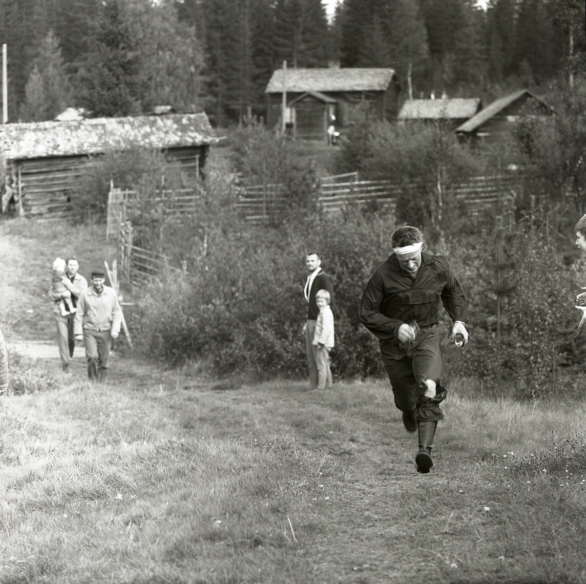 En man kommer springande upp för en backe i Svartbo 1963. Han deltar i distriksmästerskapen i orientering och en liten publik bestående av barn och tre män hejar på honom. I bakgrunden står ett fäbodlandskap med gärdesgårdar, stugor och åkermark.