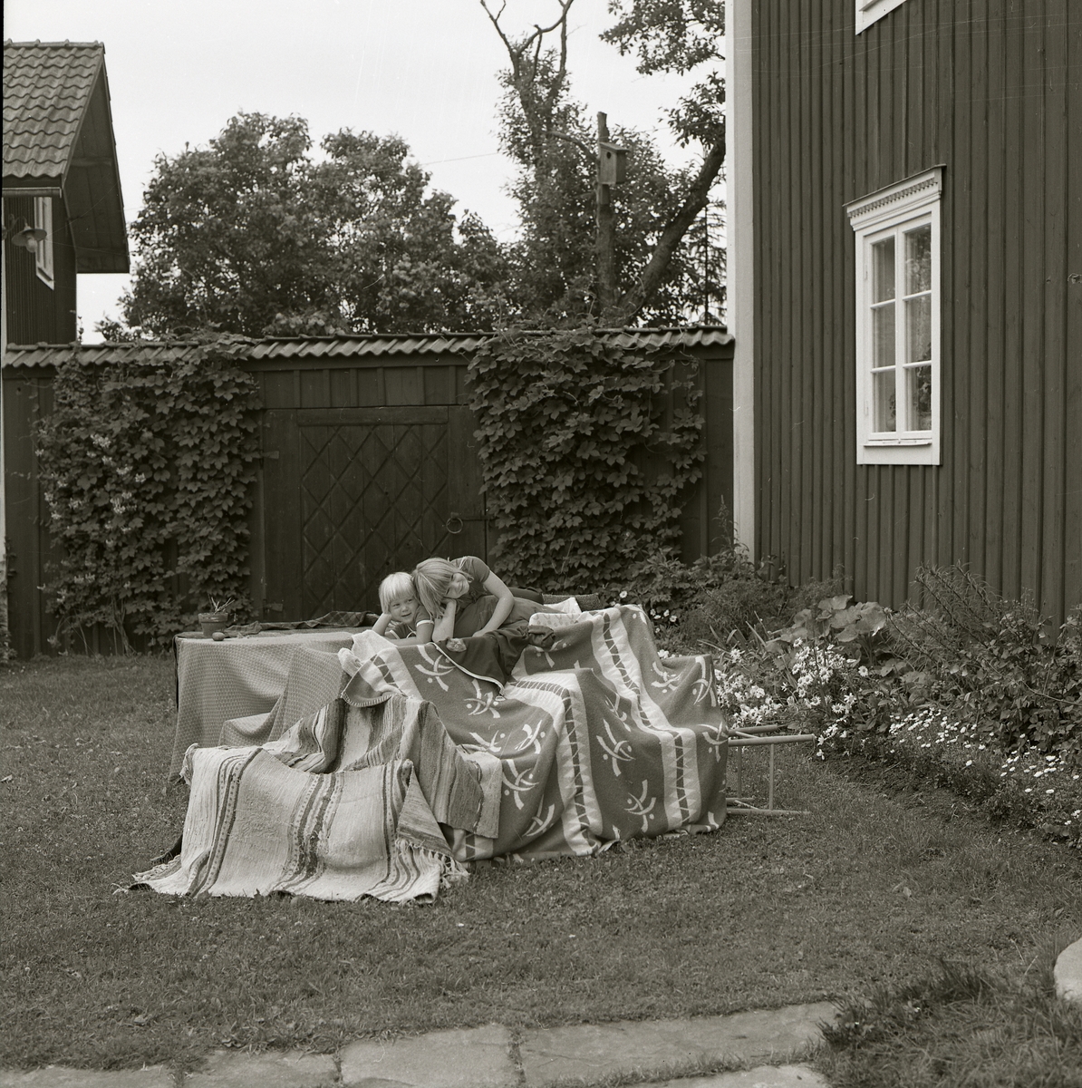 På gräsmattan framför ett hus har två flickor byggt en koja av filtar och mattor. Det är sommar 1977 på gården Sunnanåker och flickorna bär t-shirts. Bakom barnleken syns ett plank som avgränsar trädgården från lagugård och uthus.