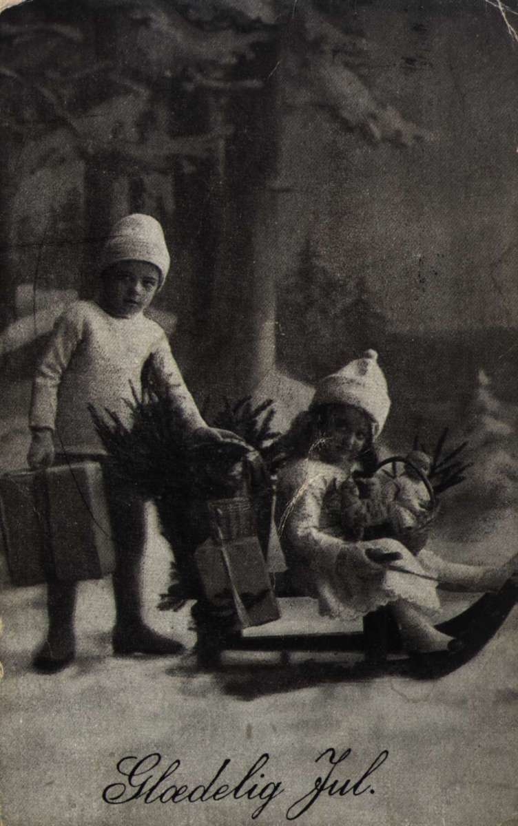 Julekort. Jule- og nyttårshilsen. Svart/hvitt fotografi. Snødekt skoglandskap. Gutt og pike med julegaver. Hun sitter på en kjelke. Stemplet 22.12.1922.
