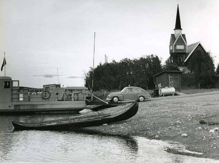 Kyrkan och färjstället i Karesuando, augusti 1961. En personbil Volvo PV är på väg att köra ombord på färjan. En träbåt ligger delvis uppdragen på stranden.