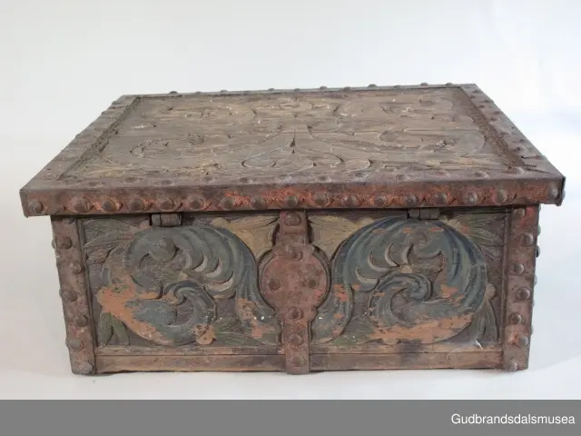 "Bladformer, smøygde band og drageornamentikk. Millom kantjarn og utskurden hev lokket fint utskorne små blad og halvrunde aggar. Vel vedlikehalde, sterk jarning og prydelege gangjarn. Truleg frå 1500 talet. Fra Haugen, Øystre Gausdal". (Jf. "Katalog for Lom Bygdesamling,1925).
Skrinrammen er holdt sammen av utvendige hjørnekantbeslag; jernhandtak i hver side. Fronten har nøkkelhullslås med beslag rundt. Lokket er satt samen av 2 bord, forsterket med jernbeslag rundt kanter på overside; to hengselbeslag festet til innside av lokk og bakstykke. Skrinsider blir holdt sammen av beslag ned langs hjørnekanter; har jernhandtak festet på jernbeslag.
Bunnen er satt sammen av 2 bord, festet til hverandre med to forsenka treplugger; bunnen var opprinnelig festet til underkant av skrinsider med 4 x 4 trenagler,seinere festet med spiker. 
Det er jernbeslag  på bakstykket midt, rundt hjørnekanter, omkring nøkkelhullslås, rundt lokk samt 2 handtak med beslag. Alle beslag er festet med store jernnagler; beslag på sidestykker er festet med små jernnagler under skrinbunnen. 3 av 8 bunnfester for beslag er fortsatt intakte.
DEKOR. De store jernnaglene med forskjøvet plassering på beslag ble sannsynligvis brukt som dekorelement; runde beslag på sider, bakstykke og front. Flatskurd på lokk med flettet, stilisert renessanseranke, der bladverk er "omgjort" til småspiraler eller rundinger.  På skrinsidene akantusskurd med c-former, bladtunger uten stilk og naturalistiske småblad i rokokkostil. Flatskurd var vanlig i Gudbrandsdalen i perioden ca. 1650-1750, mens akantusmotivet først ble vanlig på bruksgjenstander i løpet av siste halvdel av 1700-tallet. Den mørkere grønne og blå dekorfargen var vanlig i siste del på 1700-tallet; den brune bunnfargen var vanlig i bruk fra 1780-tallet. 
BRUK. Før kofferten kom i bruk hadde man reiseskrinet, som ble brukt til oppbevaring av bl.a. klær, verdisaker og mat på reiser. Skrinet hadde ofte et eller to mindre, smale rom langs den ene skrinsiden og ett større rom. De fleste reiseskrin var i tillegg forsterket med jernbeslag langs alle kanter og med en solid nøkkelhullås til sikring mot støt, slag og tyveri.