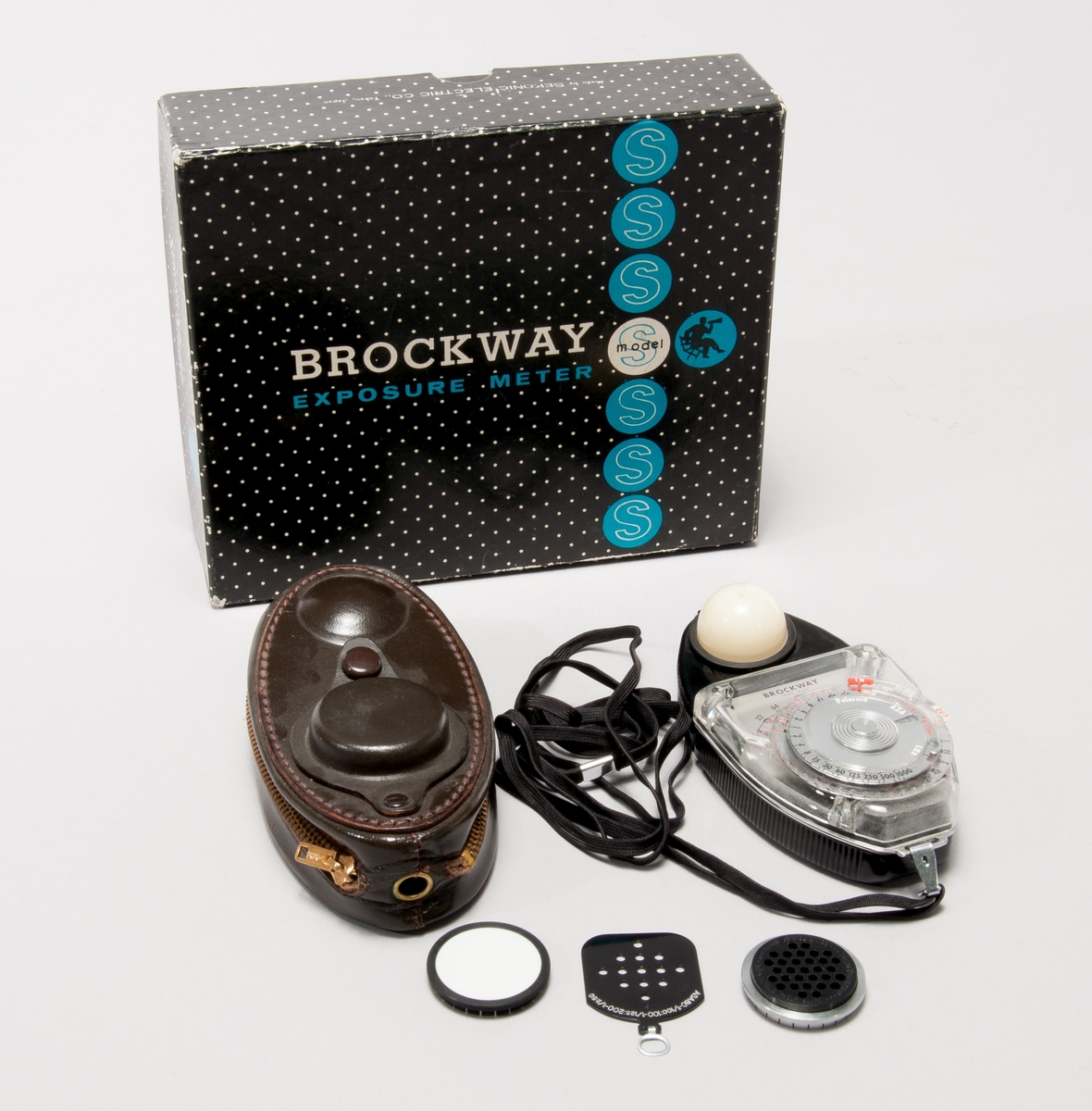 Exponeringsmätare typ Brockway, med vridbart mäthuvud. Olika bländare medföljer för justering efter filmkänslighet. Tillhörande läderetui med förvaringsmöjlighet för tillbehören.
I ask av papp.