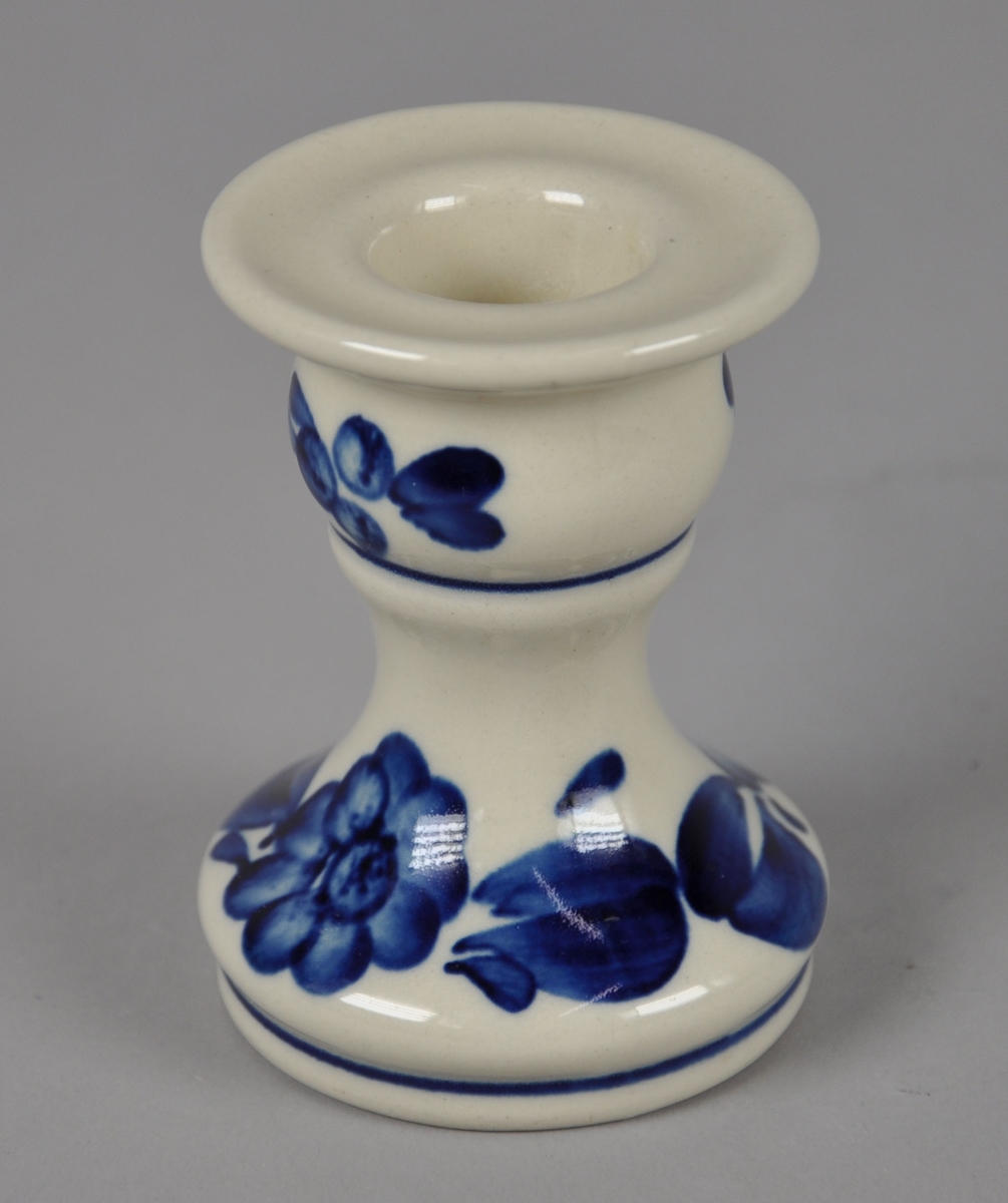 Lysestake av glassert keramikk. Har malt dekor med motiv av blå blomster, blader og bær. Har kurvet sylinderform der øverste og nederste del er utstående.