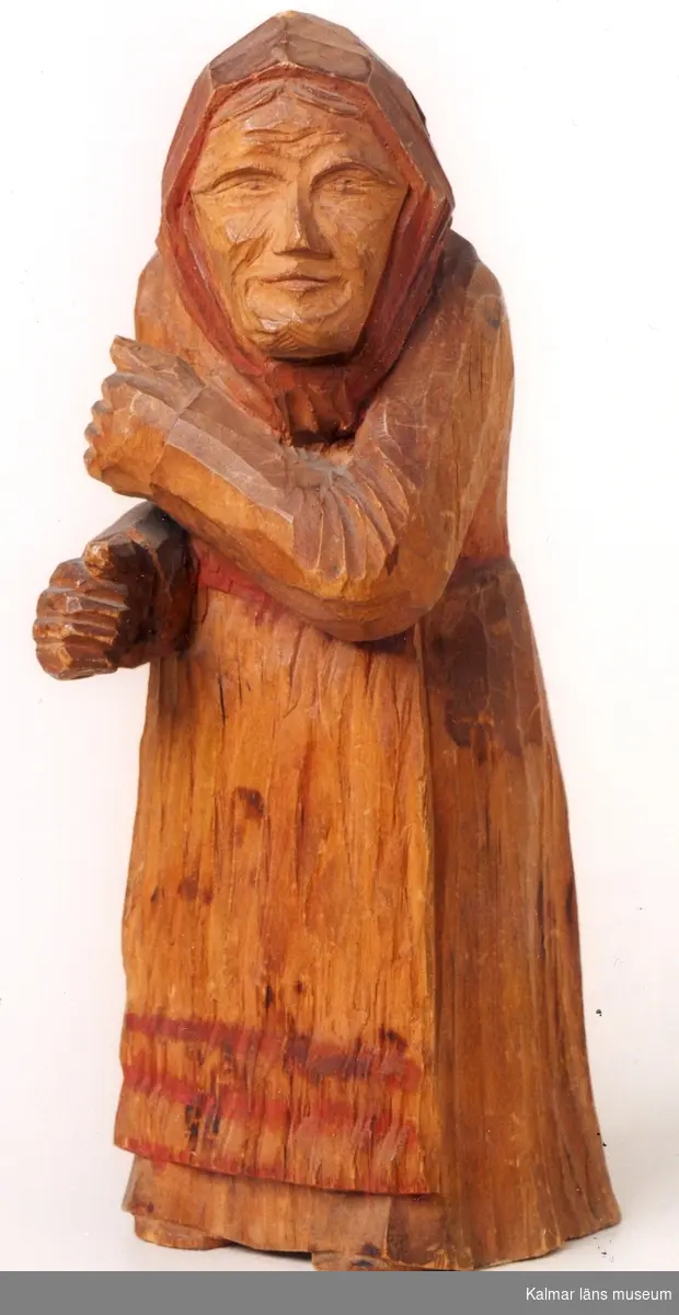 KLM 39255:5. Skulptur, av trä, delvis bemålad i rött. Gumma med sjalett och långt förkläde. Okänt, nu förkommit föremål suttit i gummans händer. Signerad, B. Ljungren, Markaryd 1918.