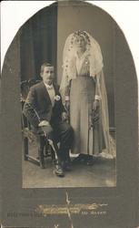 Brudebilde. Brudgommen sitter og bruden står. Hun har hvitt 