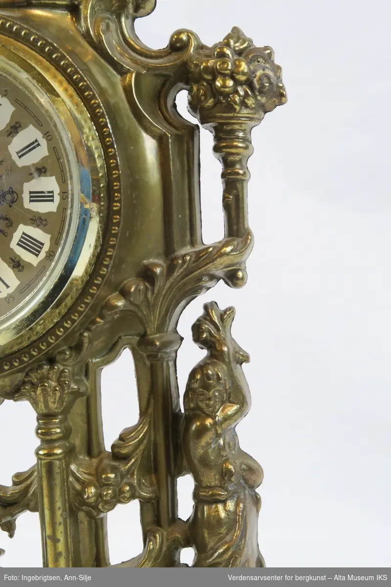 Batteridrevet ur. Stående oppsats med barokk innfatning og innfelt urskive med romertall.