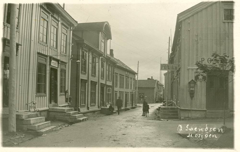 Gate med gammel trehusbebyggelse. Der er tre- fire personer i gata og der står en sykkel opp mot veggen mellom to stein trapper til venstre. På høyre side er det en butikk P.O.Wika.