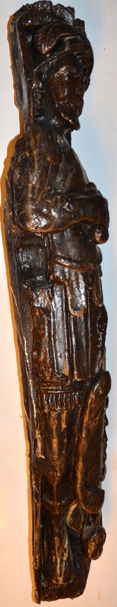 Krigare iklädd romersk rustning samt plymagerad hjälm. Mannen har långt hår och skägg. I höger hand håller mannen ett draget svärd snett över bröstet och upp mot vänster axel. Svärdsklingan är snidad separat. Vänster hand fattar om en stor, profilerad sköld. Skölden är dekorerad med en i relief skuren halv mångubbe. Skölden vilar mot vänster ben. Mannens huvud och bål är kraftigt vridna mot vänster medan underkropp och ben är placerade i en face. Höger ben är placerat framför vänster ben som till större delen är dold bakom skölden. Benen är stövelklädda. Utrymmet för fötterna är minimalt.

Figuren är placerad snett i förhållande till bakstycket. Skulpturens fotända är, sedd framifrån, i det närmaste trekantig. Skulpturens baksida visar tydliga huggspår och har ett snett urtag upptill som lämnar huvudändan nedsänkt. En markant urgröpning med rektangulärt hål kan iakttas på detta parti.

Skulpturen och bakstycket är snidat i ett stycke. Armarna är snidade separat och sitter fästade vid bålen med dymlingar. Bakstycket är profilsvängt och baksidan är slät. Nedtill är bakstycket framdraget så att det bildas en kraftig sockel på vilken figuren står. Sockeln är ornerad på sidorna. Bakstyckets övre del fortsätter snett uppåt och bildar en bred platta ovanför krigarens huvud.
Skulpturen är relativt välbevarad. Skulpturens baksida och fotända är delvist skadade.

Text in English: Warrior clad in Roman armour, with long hair and a beard. A simple plumed helmet adorns his head. The man draws his sword with his right hand across his chest and up towards his left shoulder. The blade of the sword was carved separately. The left hand grasps a large, profiled shield, decorated in relief with a profile of half the face of the moon. The shield rests against the left leg. The man''s head and body are twisted considerably to the left. The shield conceals most of the body.

The figure is placed at an angle in relation to the back-board. The foot end of the sculpture is practically triangular as seen from the front. the rear side of the sculpture has a slanted groove near the top that leaves the head end lower, indicating that the sculpture is attached partly to the outside planking.

The sculpture is carved from a single piece of wood, except for the arms which are carved separately and attached to the thorax with dowels. The figure is placed in front of a profiled back-board, with a smooth reverse side, which is drawn forward at the bottom to form a robust base upon which the figure stands. The base is simply ornamented on the sides. At the top the back-board continues above the heads to form a wide plate-like section over the warrior''s head.
The sculpture is rather well preserved.  Skulpturens baksida och fotända är delvist skadade.
