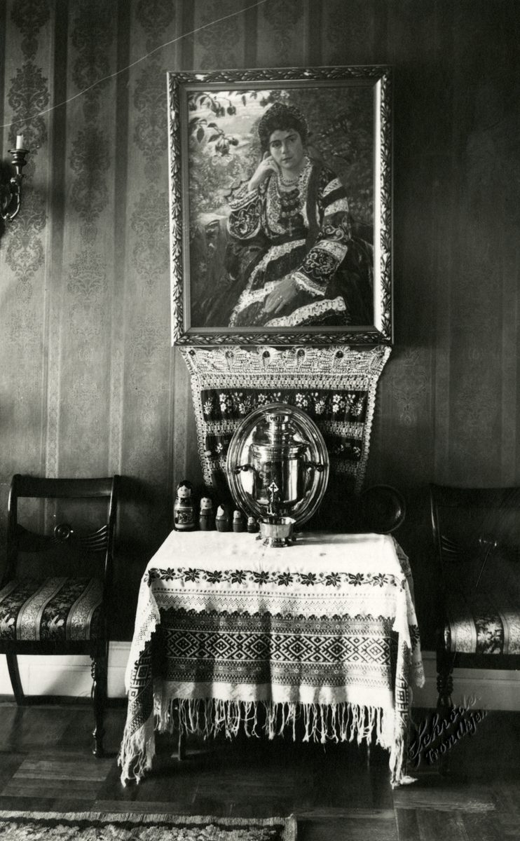 Et bilde av et portrett som forestiller Victoria Bachke i sin russiske festdrakt, malt av Alexander Piskaunoff. Under portrettet står det et bord dekket med russisk duk med korsstingsbroderier, samovar og dukker.