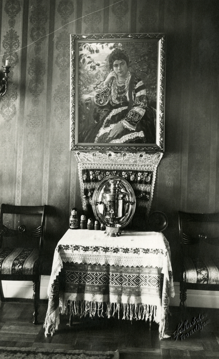 Et bilde av et portrett som forestiller Victoria Bachke i sin russiske festdrakt, malt av Alexander Piskaunoff. Under portrettet står det et bord dekket med russisk duk med korsstingsbroderier, samovar og dukker.