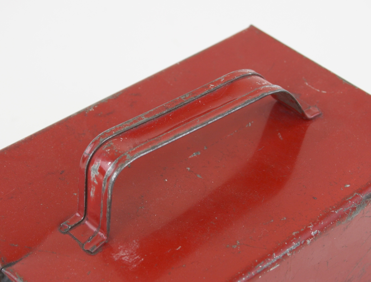 Redskap för bärplockning i rödlackerad plåt. Lådan har en lätt böjd botten med galler, ett handtag sitter på ovansidan.