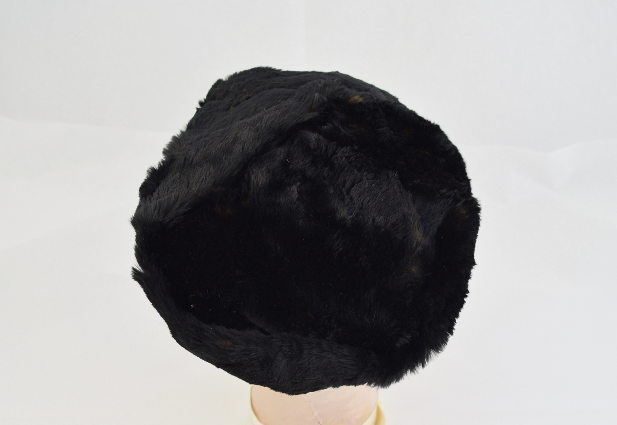 Hatt av pels med for i tynt blankt stoff. Hatten har en devis rund form og består av en rund topp med en bred kant.