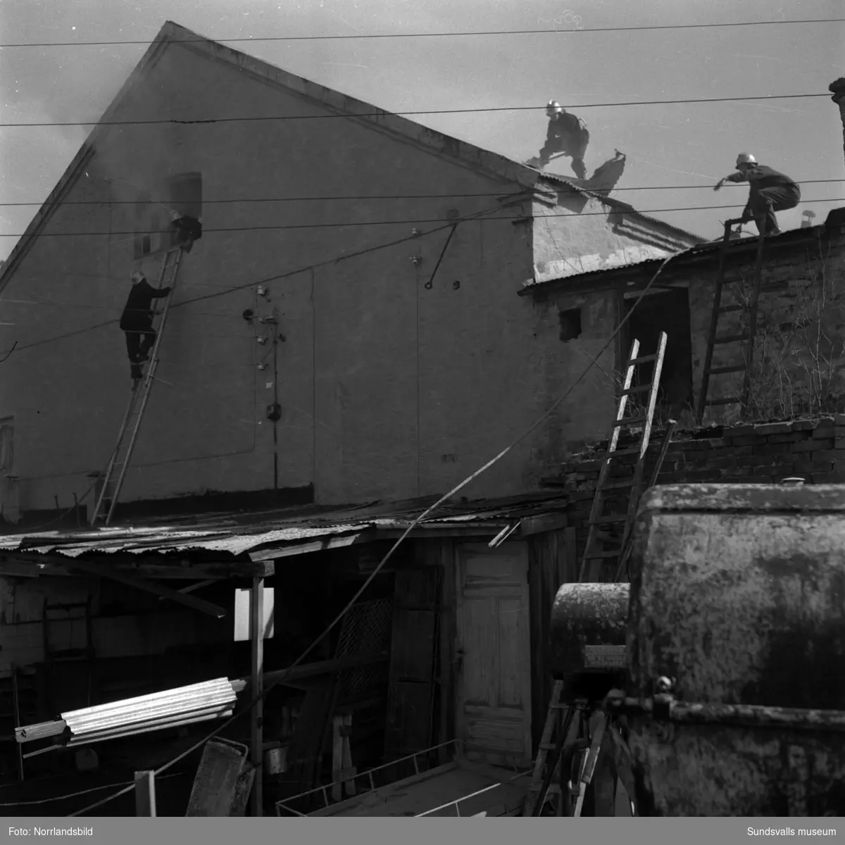 Ett par brandmän med hjälmar och yxor bryter upp taket för att bekämpa en eldsvåda i en gammal industribyggnad vid Fabriksgatan. I bakgrunden syns Västhagens sjukhus.
