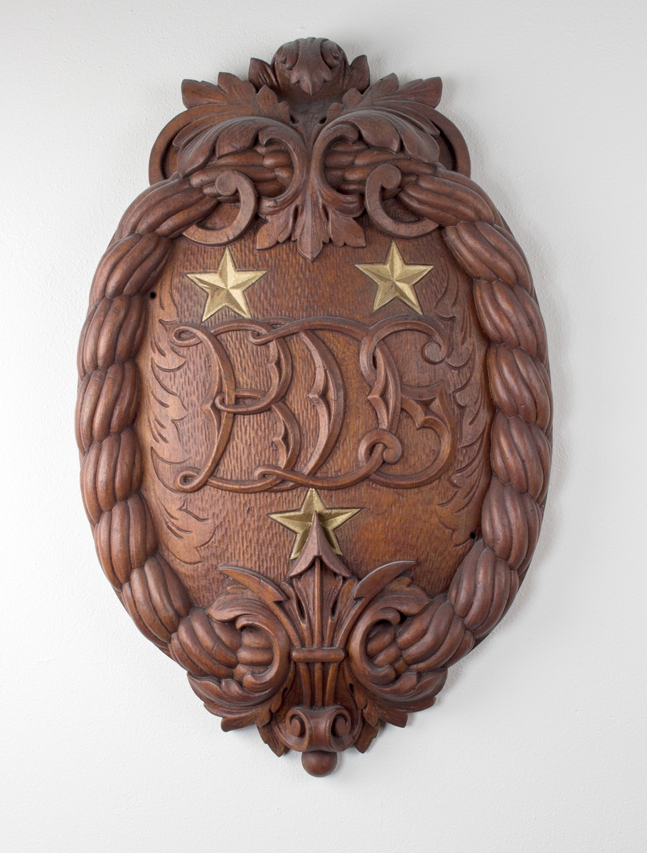 Ovalt emblem utført i tre med bokstavene BDS i senter, omkranset med tre stjerner og tau.
