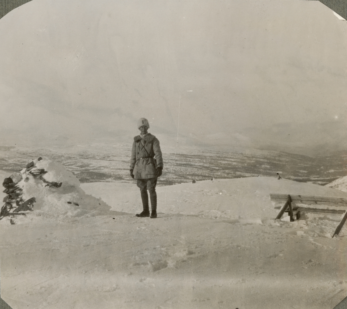 En soldat vid ett stenröse och bänk på fjället. Bakom soldaten öppnar sig ett vintrigt fjällandskap med dalgång och fjälltoppar i fjärran.