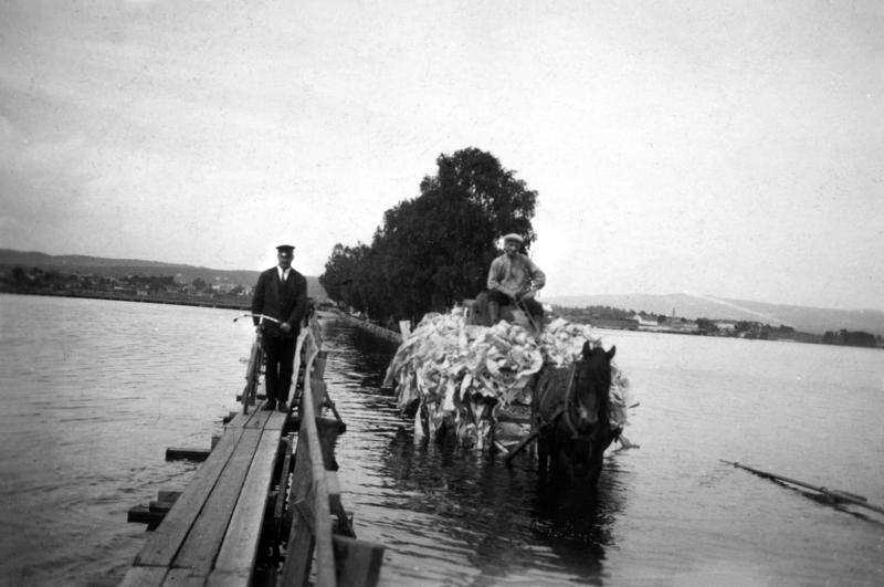 Svart-hvitt foto fra 1927. Gangbru med mann og hestetransport med stort lass som kjører på ordinær bru i flomvann. (Foto/Photo)