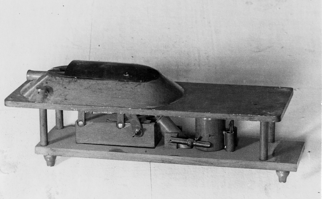 Modell av kanon föreställande en skeppskanon i tornlavettage enligt C.G. Krügers konstruktion. Sänkbar kanon med tilllhörande rekylcylinder och hydralisk pump. Allt av fernissad bok. Toppen täckt av plåtskälla. Ena rikthuven borta.