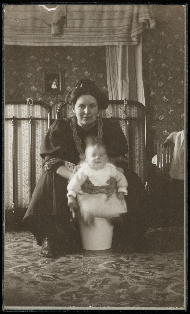 En kvinna håller i en spädbarnspojke som sitter på en hink i ett sovrum. Påskrift på baksida: "Farmor gratuleras hjärtligt på Sofiadagen 1908 af Stig med mor + far. Kortet taget den 19/4 1908."