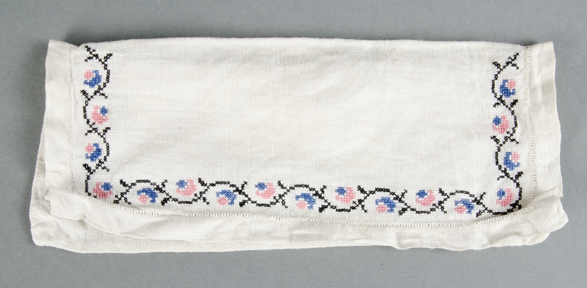 Servettväska av vit linnelärft, handsydd med hålsöm på locket och blombård i korsstygn i svart, rosa och blått. 
Ostruken och skrynklig.

