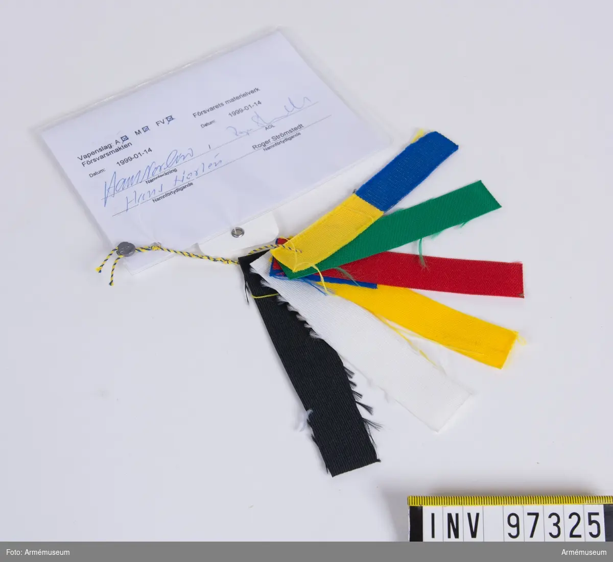 Knippe med prov på band i olika färger. Varje färg har eget M-nummer. På den vidhängande modellappen anges alla M-nummer samt att teknisk bestämmelse finns i avtalet. Lappen är underskriven 1999-01-14 av Hans Norlén och Roger Strömstedt.