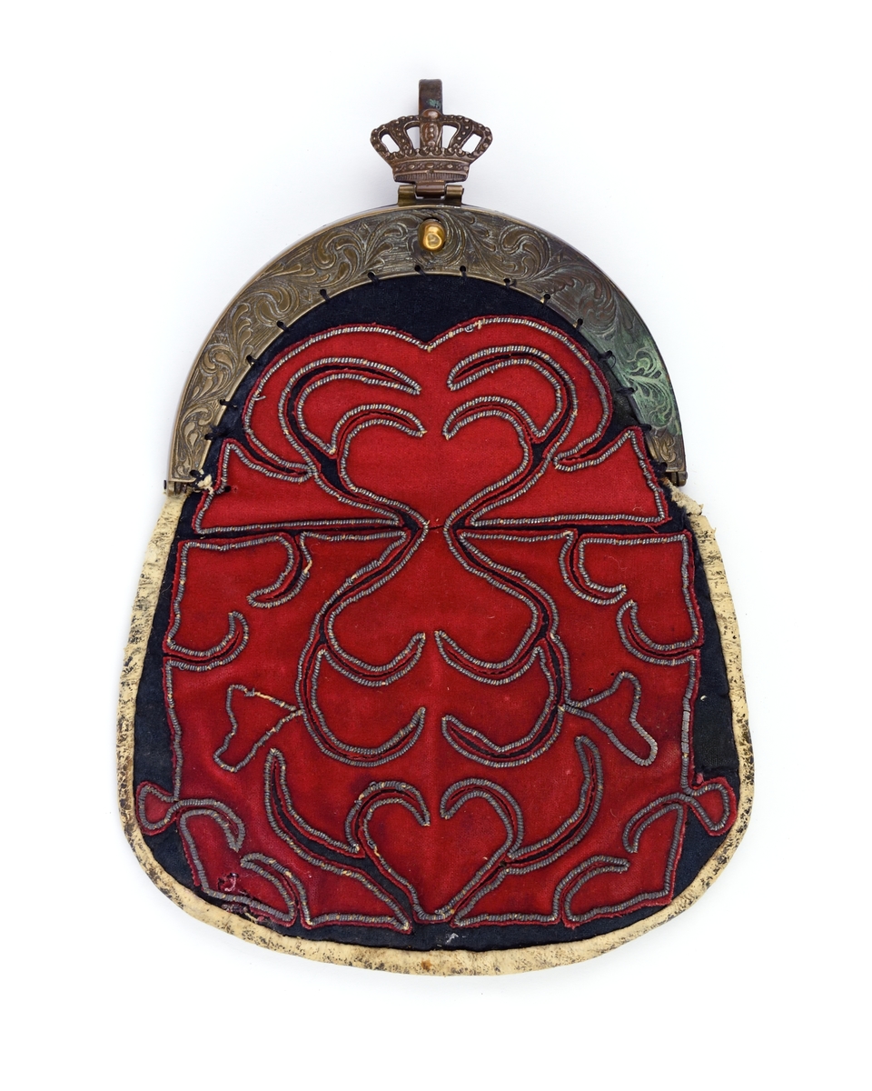 Kjolväska med applikationer i rött, kantat med tenntråd på svart botten. Väskan har skinn på baksidan och är fodrad med rutigt linnetyg. På bygeln sitter en krona.