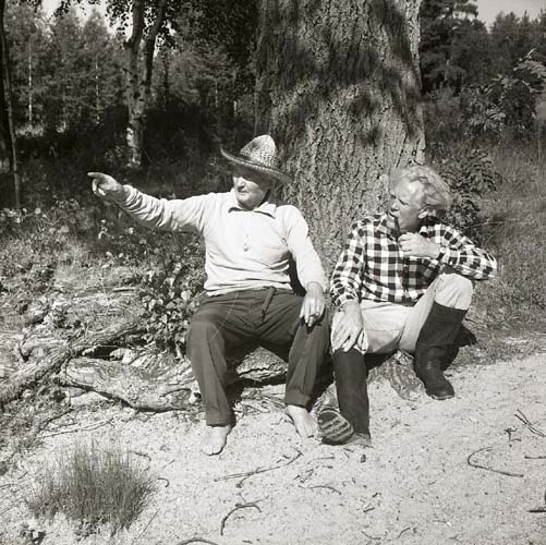 Två män sitter lutade mot en trädstam vid Ängratörn 19 juli 1966. Ena mannen pekar mot något och den andra tittar mot det hållet medan han tuggar på något han håller i handen.