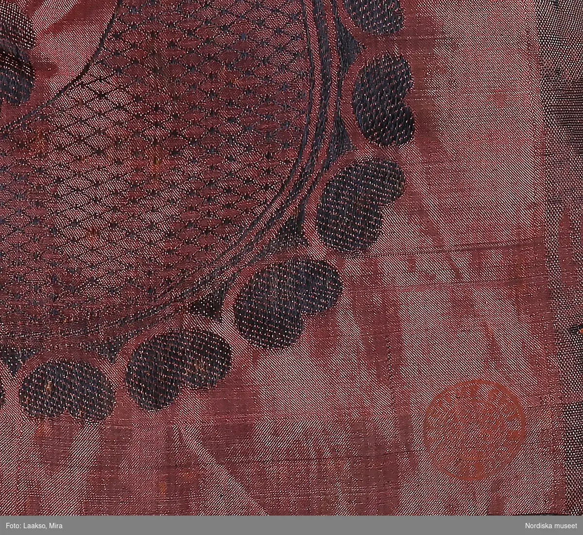 Kvadratiskt halskläde av sidentaft i svart och rödrosa.  Mittspegeln mörk med jacquardvävt mönster: rutindelning med ornamenterade bårder, i varje ruta en  svängd blomkvist. Bred kantbård med  stora vågböjda gallerband med mellanliggande blommor. Handsydda fållar, tvåknuten krusad frans runtom av tvinnat silke i rosa och svart. I ena hörnet en röd rund stämpel med texten: Stockh. hallstemp. 1849.
Buren som brudschal omkr. 1850.
Anm. I fint skick, en mindre blekt fläck.
/Berit Eldvik 2011-12-07