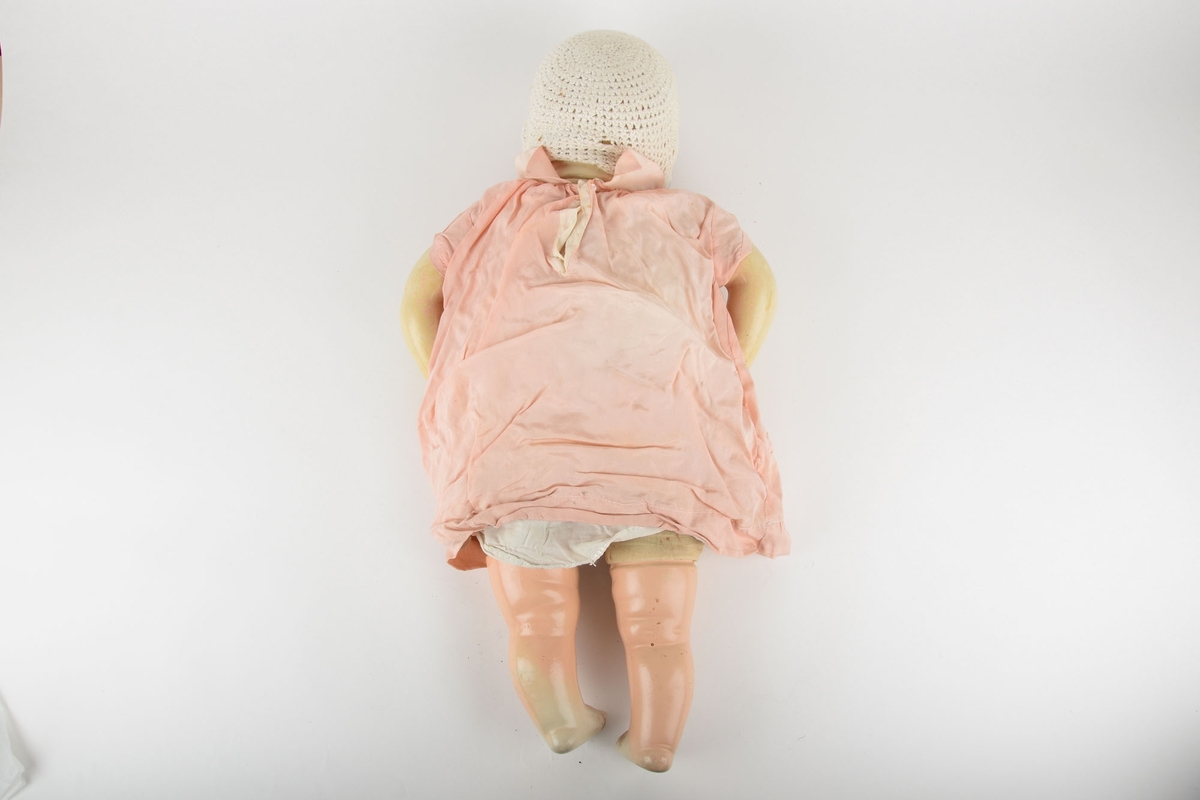 Babydukke med tøykropp. Hode, armer og bein i plastlignende materiale. På hodet har dukken strikket lue med knytting under haken. Kledd i hvit underkjole, med lyserosa kjole over, som har enkel brodering på hver side.