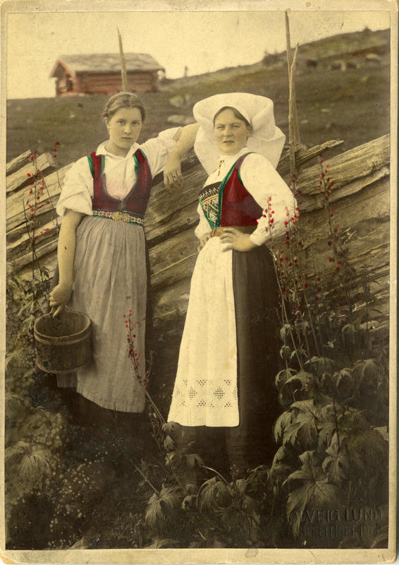 Kolorert fotografi av to kvinner med Hardangerdrakt foran et gjerde på seteren. 1908. (Foto/Photo)