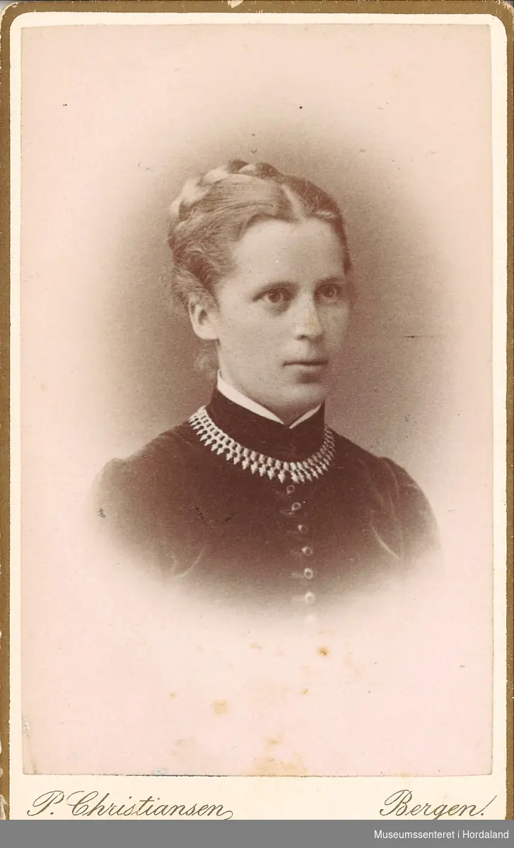 portrettfotografi av yngre kvinne med blondt hår fletta og oppsett, mørk fløyelskjole med mange små knappar og bord rundt halsen