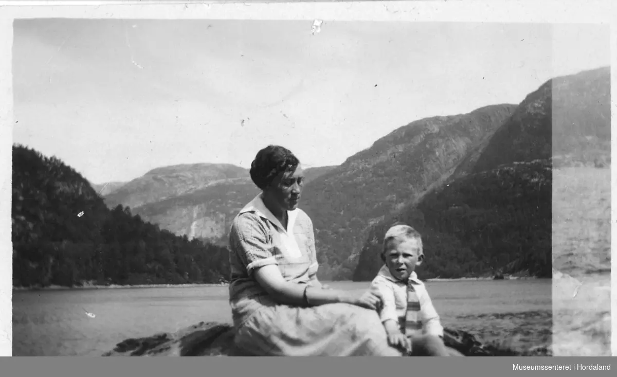 amatørfotogafi av kvinne med ein liten gut som sit i vannkanten med fjord og fjell bak seg
