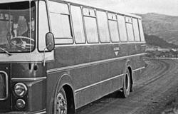 Buss. Kjører på grusvei med fjell i bakgrunnen.